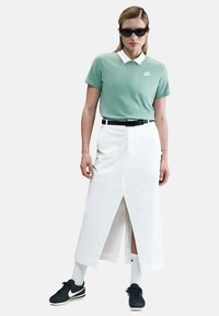 Femme portant un polo Nike vert menthe, une jupe blanche taille haute avec fente à l'avant, une ceinture noire, des baskets Nike noires, des chaussettes blanches et des lunettes de soleil noires.