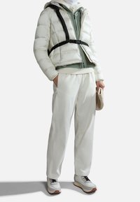 Biała puchowa kurtka z czarnym uprzężą, nałożona na zieloną bluzę z kapturem. W połączeniu z białymi spodniami i masywnymi białymi sneakersami, trzymająca beżową torbę.