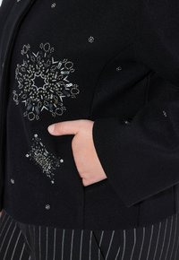 Main dans la poche d'une veste noire décorée de motifs floraux en perles et paillettes argentées, associée à un pantalon noir à rayures.