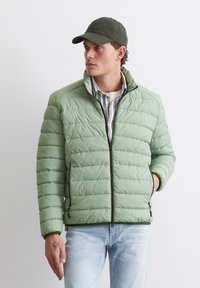 Ljusgrön puffjacka med quiltad textur, svarta dragkedjor och ståkrage. Bärs över en randig skjorta och matchas med ljusa jeans.