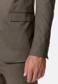 Blazer a scacchi marrone con tasca dritta, vestibilità su misura e polsino abbottonato. Tessuto dalla texture liscia e cuciture a contrasto sottili.