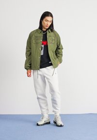 Olijfgroene jas met geborduurd ontwerp, zwart graphic t-shirt, witte sweatpants en grijze sneakers met zwarte accenten, staand op een blauwe ondergrond.