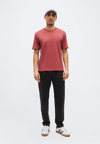 Mann trägt eine hellgraue Cap, staubrotes kurzärmeliges T-Shirt, schwarze Hose und weiße Turnschuhe, steht vor einem schlichten weißen Hintergrund.