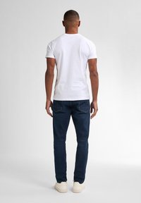 Camiseta blanca de algodón, de manga corta, combinada con pantalones vaqueros oscuros de corte ajustado y calzado blanco. El fondo es neutro y discreto.