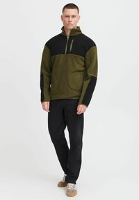 Fleece-pullover i oliven grønn og svart, med glidelås i nakken, to frontlommer og kontrasterende ermeinnsatser. Glatt tekstur, avslappet passform.