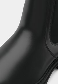 Gros plan d'une botte cheville en cuir noir avec panneau latéral élastique et coutures visibles le long de la semelle et du bord supérieur.