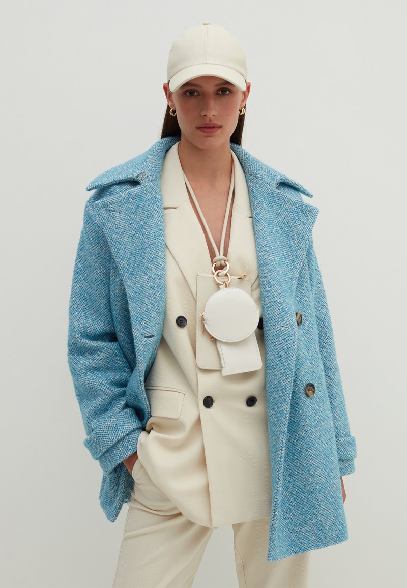 Blauer Tweed-Mantel mit übergroßem Kragen, cremefarbener Blazer und Hose. Eine cremefarbene Mütze und eine kleine, runde Umhängetasche mit goldenen Akzenten sind enthalten.