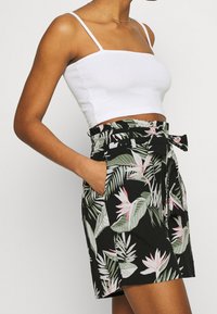 Bílý žebrovaný crop top s tenkými ramínky společně s černými šortkami s tropickým vzorem listů a květin; vysoký pas s mašlí.