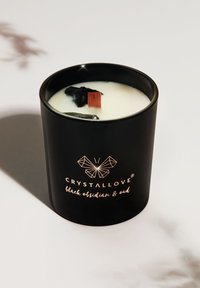 Candela nera in un contenitore nero lucido, con interno in cera bianca, piccoli cristalli neri e un accento in legno marrone sulla parte superiore. Logo dorato visibile.