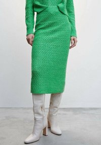 Grünes gestricktes Zweiteiler-Outfit bestehend aus einem strukturierten Pullover und einem knielangen Rock, kombiniert mit weißen kniehohen Absatzstiefeln.
