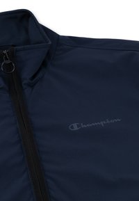 Navy windbreaker jacket with a zip closure, ribbed collar, and subtle "Champion" logo printed on the chest. Smooth texture, lightweight material.