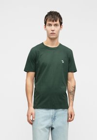 Dark green cotton t-shirt featuring a round neck, short sleeves, and a small embroidered logo on the left chest. Worn with light blue jeans.