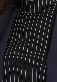Černá tkanina s bílými vertikálními pruhy, s vysokým límcem. Design obsahuje jednobarevnou námořnickou vrstvu, která vytváří vrstvený vzhled.