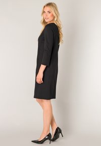 Zwarte nauwsluitende jurk met driekwart mouwen, gladde textuur, knielengte, gecombineerd met glanzende zwarte hoge hakken.