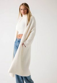 Lange witte gebreide cardigan met een open voorkant, gestructureerde stof en lange mouwen, gecombineerd met een witte cropped trui en lichtblauwe spijkerbroek.
