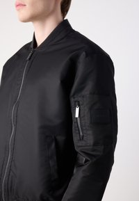 Veste bomber noire à finition lisse, poche latérale zippée et petit patch sur la manche avec le nom de la marque.