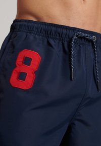 Pantaloni da bagno da uomo di colore blu navy con vita elasticizzata, chiusura con lacci e un evidente numero 8 ricamato in rosso sul lato sinistro.
