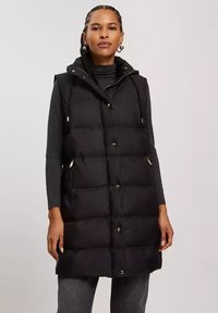 Czarna kamizelka puchowa z kapturem, z zamkiem błyskawicznym i zapięciem na napy, bocznymi kieszeniami oraz pikowaną fakturą. Wytrzymały materiał, sięgająca do kolan konstrukcja.