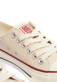 Beige lõuendist tennisekingad punase logo sildiga, metallist silmadega ja kreemjatel paeladel. Omab valget kummist tallat, millel on punane triip aktsent.