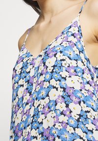 Kwiatowy top na ramiączkach w kolorach niebieskim, fioletowym i białym. Posiada dekolt w kształcie litery V oraz cienkie ramiączka, wykonany z lekkiego materiału z wzorem kwiatowym.