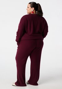 Vrouw met donker haar in een paardenstaart, gekleed in een bordeauxrode geribde trui en broekset met witte spijkerschoenen, glimlachend en naar opzij kijkend.