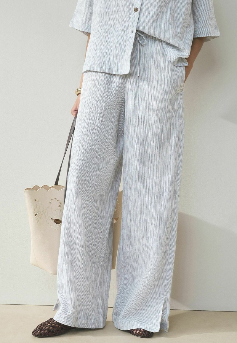 Personne portant un pantalon large texturé gris clair et un haut assorti, tenant un sac fourre-tout beige avec découpes et portant des chaussures tissées marron foncé.
