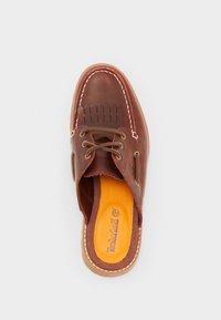 Zapatilla de cuero marrón tipo slip-on con detalles de costura, suela beige y plantilla naranja con logotipo. Punta redondeada y diseño superior abierto.