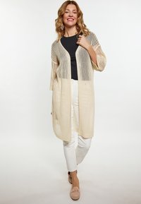Cardigan lungo leggero e a maglia aperta di colore crema con maniche a tre quarti, indossato su un top nero e pantaloni bianchi, caratterizzato da una vestibilità comoda.