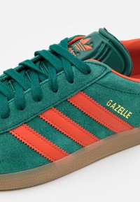 Zelene gamuzne tenisice s narančastim kožnim detaljima, s tri pruge i oznakom "GAZELLE" u zlatnoj boji sa strane, te gumenim džonom.
