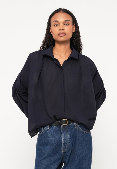Camicia oversize blu navy con colletto a punta e maniche arricciate, abbinata a jeans blu a vita alta e una cintura nera. Texture liscia, dettagli minimi.