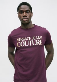 Jauns vīrietis ar pieguļošu bordo T-kreklu ar zelta uzrakstu "Versace Jeans Couture" uz vienkārša baltā fona.