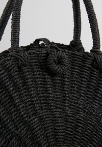 Svart vävd väska med en texturerad rund kropp, tjocka tvinnade handtag och en knuten accentdetalj högst upp, tillverkad av naturliga fibrer.