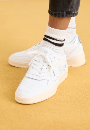 Zapatillas blancas con una parte superior de cuero suave, puntera perforada, cordones planos y una suela acolchada. Combinadas con calcetines blancos de canalé que tienen rayas negras.