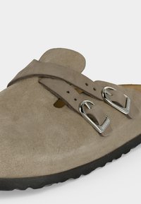 Μπεζ σουέτ παπούτσι τύπου slip-on με δύο ρυθμιζόμενες ασημένιες μεταλλικές αγκράφες και μαύρη υφή σόλα.