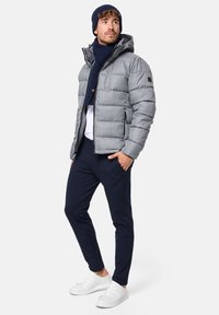 Grå quiltet pufferjakke med hætte, Navy tørklæde, hvid T-shirt, navy bukser og hvide sneakers. Har en glat tekstur og lynlås-lommer.