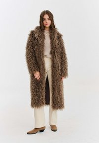 Dlouhý hnědý kabát z umělé kožešiny s vycpávanou strukturou, otevřeným předním dílem a dvěma bočními kapsami, nošený přes krémový svetr a béžové kalhoty.
