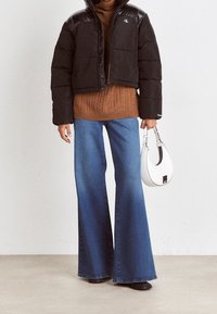Giacca a vento nera corta con dettagli lucidi, maglione a trecce marrone, jeans blu con gamba svasata e borsa bianca con un accento nero.
