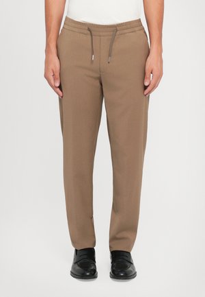 Brune tapered joggebukser med elastisk midjebånd og justerbar snøring, med en glatt tekstur og sidelommer. Bært med svarte sko.
