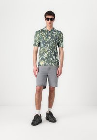 Kortermet skjorte med krage i grønn med et hvitt og gult abstrakt mønster, kombinert med grå denimshorts og svarte sneakers.