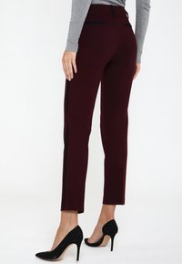 Pantalons ajustés bordeaux avec des rayures latérales noires, fabriqués à partir d'un tissu lisse. Ils présentent une taille mi-haute et des poches arrière, associés à des escarpins noirs.