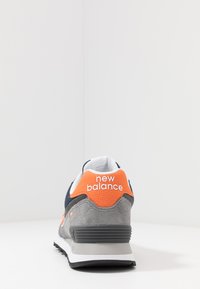 Sneakers cu un superior din suede gri, cu accente de plasă albastru închis, logo portocaliu și o talpă moale gri. Vedere din spate.