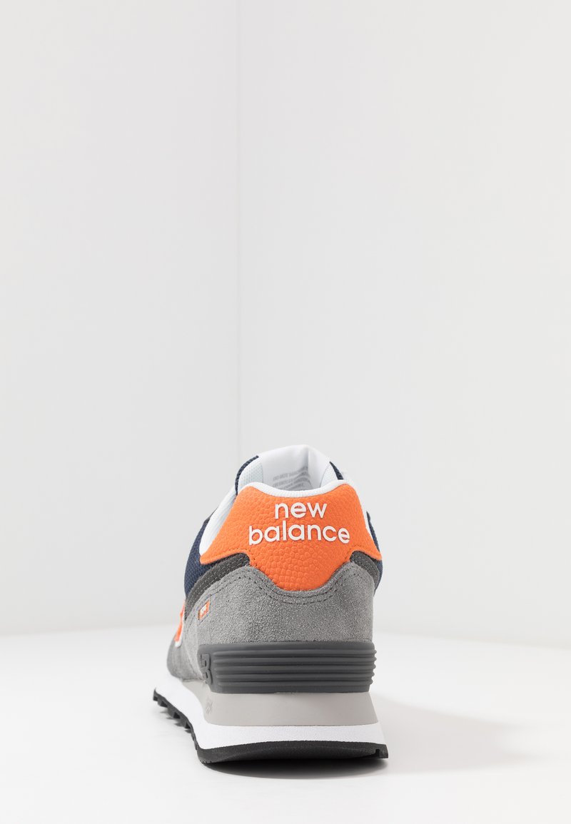 Sneaker met een grijze suède bovenkant en marineblauwe mesh accenten, oranje logo en een gewatteerde grijze zool. Beeld vanaf de achterkant.