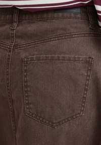 Gros plan sur la poche arrière d'un pantalon en jean marron avec des coutures détaillées et une partie d'une chemise rayée bordeaux et blanche au-dessus.