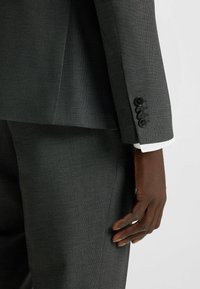 Grau strukturierte Anzugjacke mit tailliertem Design. Verfügt über vier schwarze Knöpfe am Ärmel und einen kontrastierenden weißen Hemdärmel, der sichtbar darunter hervorschaut.