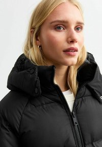 Chaqueta acolchada negra con diseño acolchado, cuello alto y cierre de cremallera; cuenta con botones circulares y una textura suave.