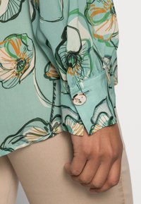 Primer plano de una mano con pantalones beige y una blusa ligera azul floral con puños abotonados que presenta patrones de flores en naranja y verde.