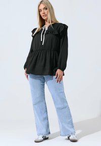 Blogin nainen seisoo päällä musta pitkähihaisen röyhelöpaidan, vaaleansinisten leveälahkeisten farkkujen ja valkoisten tennareiden, joissa on mustia raitoja.