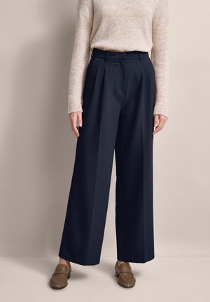 Marinefarbene weite Hosen mit Falten und tailliertem Bund. Kombiniert mit einem cremefarbenen Strickpullover und braunen Loafers, vor einem neutralen Hintergrund.