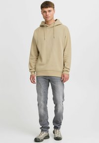 Beige kapucnis pulóver zsinórral, világosszürke farmernadrág felett viselve. Kiemelkedő részletei közé tartozik a laza szabás és a texturált anyag.