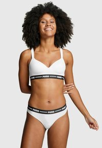 Weißer Sport-BH mit verstellbaren Trägern und einem schwarzen elastischen Band mit Logo. Passende weiße Bikini-Unterteile mit einem schwarzen Bund. Glatte Textur.